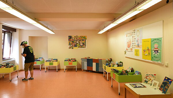 Das Kinderzimmer der Bibliothek mit vielen bunten Büchertrögen mit Bilderbüchern für die ganz Kleinen. Ein Mann stöbert in einem Trog.