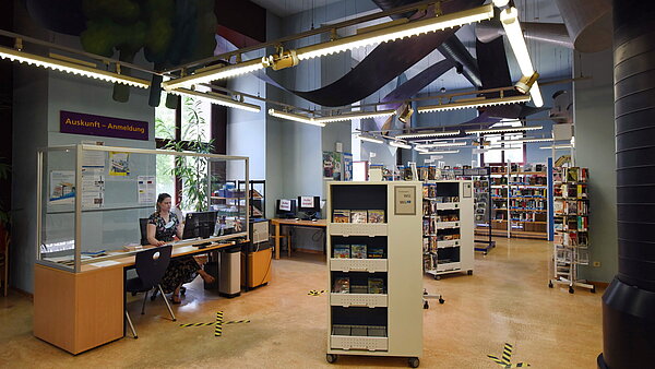Der zweite große Raum aus einer anderen Perspektive. Er hat auch sehr hohe Decken. Links ist die Auskunft zu sehen. Am Tisch sitzt eine Mitarbeiterin hinter einer Plexiglasscheibe. Im Raum stehen viele Bücherregale