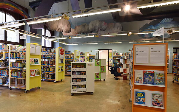 Die Kinder- und Jugendbibliothek von innen. Viele bunte Bücherregale sind zu sehen. Eine Frau sitzt in der Hocke vor einem davon und stöbert