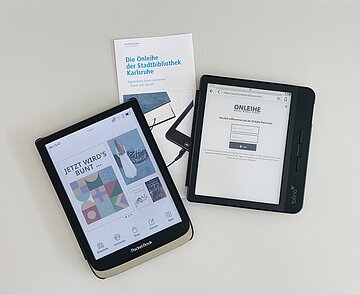 Ein eingeschaltetes Pocketbook, ein Tolino, der die Anmeldemaske der Onleihe zeigt, und ein Flyer mit der Aufschrift "Die Onleihe der Stadtbibliothek Karlsruhe". Alles auf einem hellen Tisch liegend.
