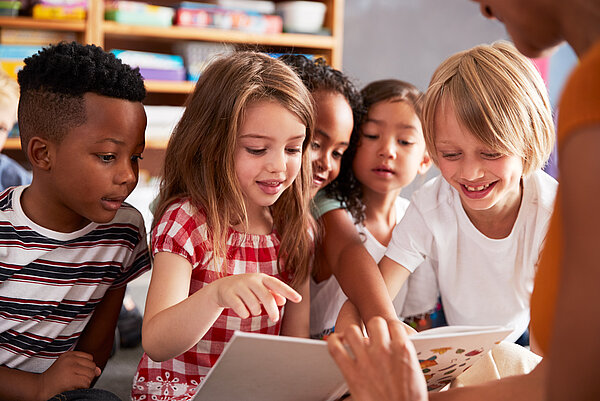 Eine Gruppe kleiner Kinder sitzt auf dem Boden, während ihnen ein Buch von einer Frau vorgelesen wird. Sie schauen fröhlich auf das Buch.