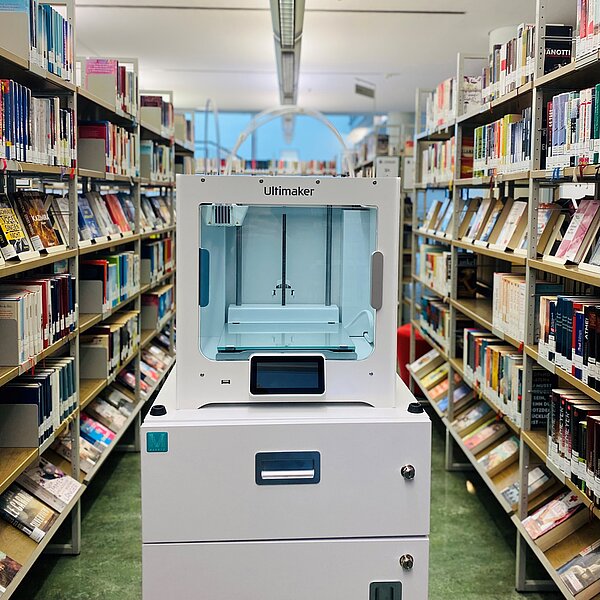 Der 3D-Drucker steht inmitten der Bibliothek zwischen Bücherregalen