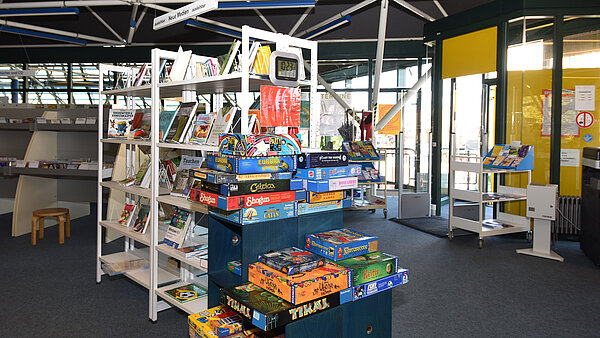 Innenansicht der Stadtteilbibliothek Nereut. Das volle Bücherregal "Neue Medien" und gestapelte Brettspiele sind zu sehen.