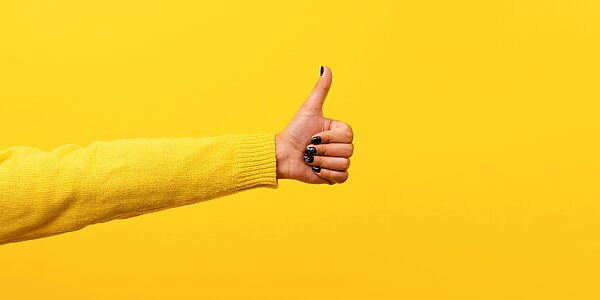 Ein ausgestreckter Arm der von links ins Bild ragt und den Daumen hoch zeigt, vor gelbem Hintergrund mit gelbem Longsleeve.