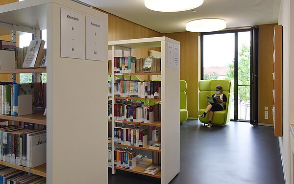 Ein weiterer Raum in der oberen Etage ist abgebildet. Hier sind wieder zwei Regale mit Büchern zu sehen und im Hintergrund ein großer grüner Sessel vor einem großen Fenster. Auf dem Sessel sitzt eine Leserin