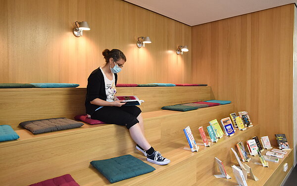 Eine Frau sitzt mit einem Buch in der Hand auf einem Sitzkissen auf der Lesetreppe. Hinter ihr an der Wand hängen Lampen die ein gemütliches Licht machen. Auf der Treppe sind Bücher ausgestellt.