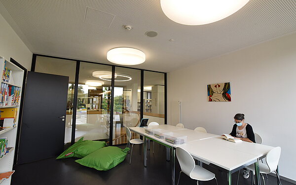 Im Lernstudio in der oberen Etage stehen große weiße Tische und Stühle. An der Decke hängen große runde Leuchten. Am Tisch sitzt eine Frau und schaut in ein Buch. Auf dem Boden befinden sich grüne große Sitzsäcke