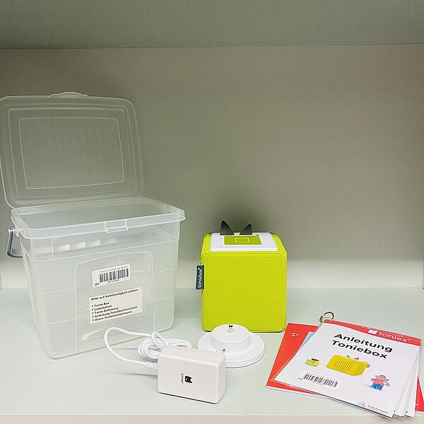 Auf dem Bild ist die druchsichtige Plastikbox zu sehen in der die Toniebox ausgeliehen wird. Daneben steht eine grüne Toniebox, davor liegt die Ladestation und daneben die Anleitung zur Benutzung der Toniebox.