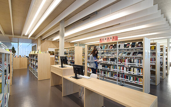 Viele Bücherregale sind zu sehen. Im Vordergrund Tische aus hellem Holz mit PC-Bildschirmen. An einem Bücherregal steht eine Frau und schaut in ein Buch