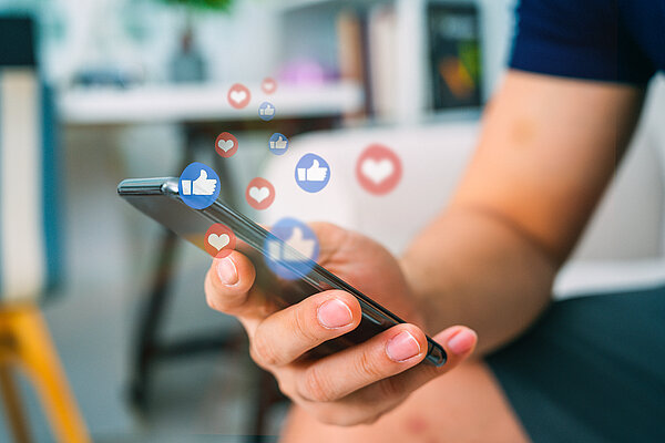 Eine Nahaufnahme von einer Hand die ein Smartphone hält, darüber steigen kleine Like-Daumen und Herzen in die Luft 
