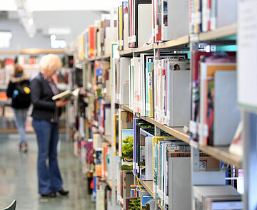 Eine Erwachsene am Bücherregal. Das regal ist im Vordergrund scharf, die Person im Hintergrund unscharf