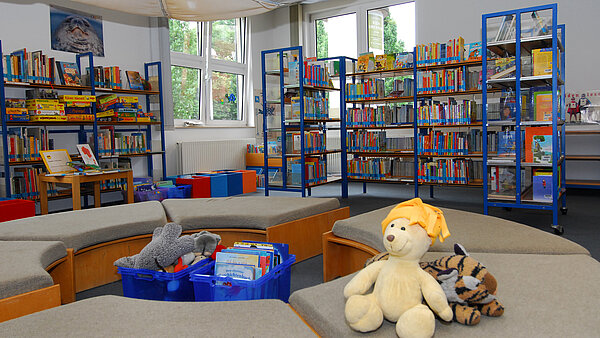 Im Vordergrund sind Sitzmöbel zu sehen, die einen Kreis ergeben und auf denen zwei Kuscheltiere liegen. In der Mitte befinden sich Boxen mit Kinderbüchern und Kuscheltieren. Im Hintergrund volle Bücherregale mit Kinderbüchern